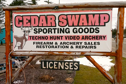 Sign for Cedar Swamp Sporting Goods in the Petoskey Area - bold orange and black lettering image of a buck is on the sign as well - small sign rotating "licences"