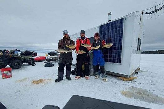 three men in warm weather gear standing side by side each holding two fish out in front of them. They are all standing in front of a shelter that has been placed on a frozen lake