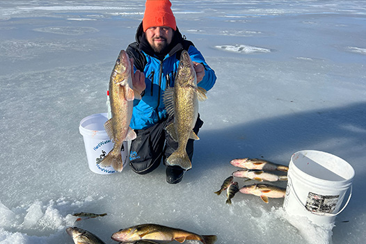 man in orange knit hat kneeling on a frozen lake holding two fish with other caught fish laying on one side of him and in front of him of a variety of sizes 